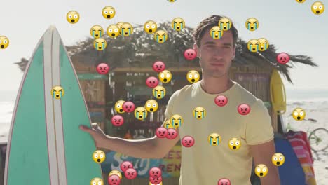 Emojis-Mit-Mehreren-Gesichtern-Schweben-Vor-Dem-Porträt-Eines-Kaukasischen-Mannes-Mit-Surfbrett,-Der-Am-Strand-Lächelt