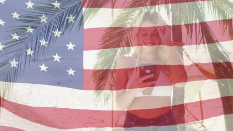 Animation-of-american-flag-waving-over-smiling-woman-using-smartphone-on-beach