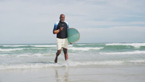 African-american-senior-man-walking-on-a-beach-holding-surfboard-and-running-out-of-the-sea
