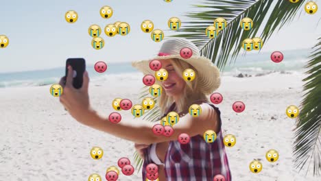 Multiple-face-emojis-floating-against-caucasian-woman-taking-a-selfie-with-smartphone-on-the-beach