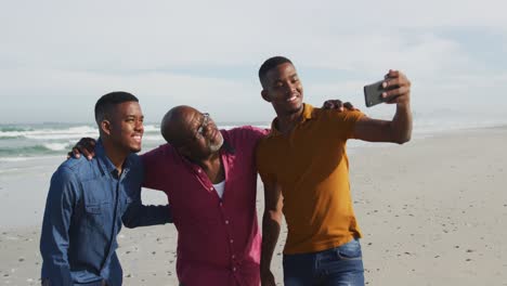 African-american-senior-father-and-twin-teenage-sons-standing-on-a-beach-using-smartphone