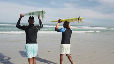 African-american-father-and-teenage-son-standing-on-beach-holding-surfboards-on-heads-and-talking