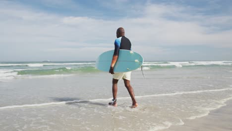 African-american-senior-man-walking-on-a-beach-holding-surfboard-and-running-into-the-sea