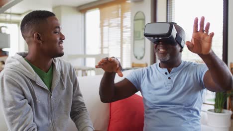 African-american-senior-father-sitting-on-a-couch-using-a-vr-headset-with-his-teenage-son-smiling