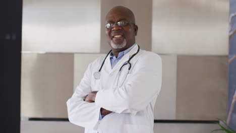 Lächelnder-Afroamerikanischer-älterer-Männlicher-Arzt-In-Weiß,-Der-Die-Arme-Verschränkt-Und-In-Die-Kamera-Blickt