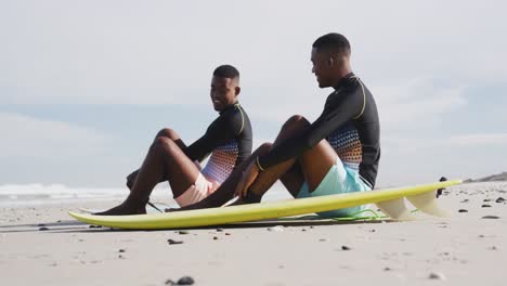Happy-african-american-teenage-twin-brothers-sitting-by-surfboards-on-a-beach-talking