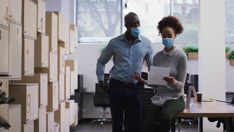 Diverse-male-and-female-business-colleagues-in-face-masks-talking,-woman-holding-document-in-office