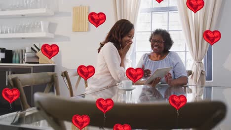 Animation-of-heart-icons-over-two-women-talking-and-using-tablet-at-home