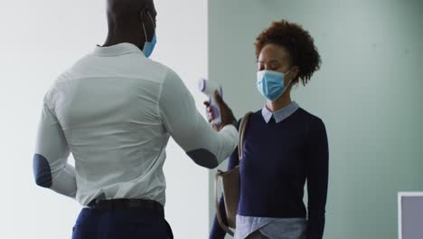 African-american-businessman-in-face-mask-disinfecting-hands-of-female-colleague-arriving-at-office