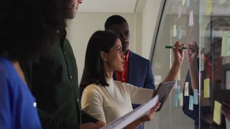 Diverse-group-of-colleagues-in-meeting-room-mixed-race-businesswoman-writing-notes-on-glass-wall