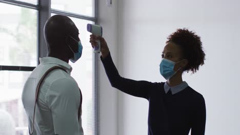 African-american-businessman-in-face-mask-having-temperature-taken-by-colleague-on-arrival-at-office
