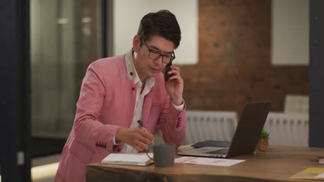 Elegant-Gekleideter-Asiatischer-Geschäftsmann,-Der-Mit-Laptop-Am-Schreibtisch-Sitzt-Und-Sich-Notizen-Macht