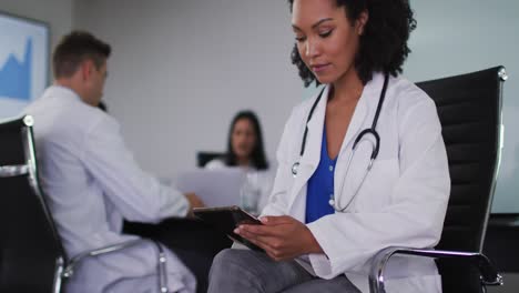 Mixed-race-female-doctor-sitting-in-meeting-room-using-tablet-looking-to-camera-smiling