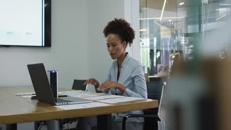 Mixed-race-businesswoman-sitting-at-desk,-checking-documents-and-using-laptop-in-office