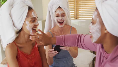 Diverse-group-of-happy-female-friends-with-towels-on-heads-and-cleansing-masks-taking-selfie-at-home