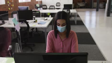 Mixed-race-businesswoman-wearing-face-mask-sitting-at-desk-using-computer