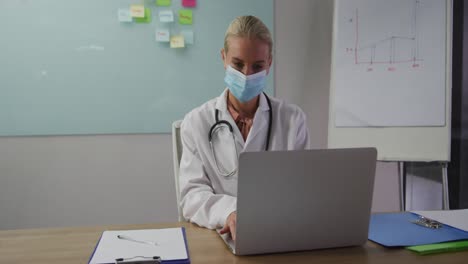 Caucasian-female-doctor-wearing-mask-sitting-at-desk-in-meeting-room-using-laptop