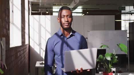 African-american-businessman-holding-laptop-looking-ahead-shifting-gaze-to-camera-and-smiling