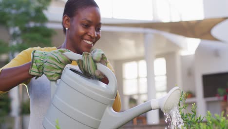 Happy-african-amercian-woman-wearing-gloves-watering-flowers-in-garden