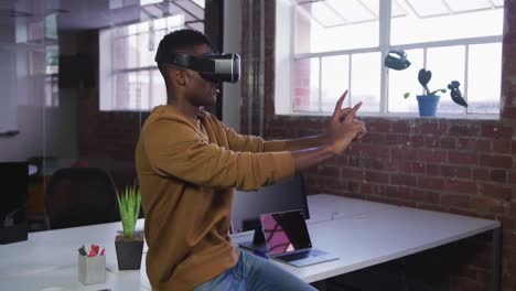 African-american-businessman-sitting-on-desk-using-vr-headset-and-gesturing