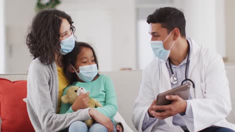 Hispanic-male-doctor-talking-to-mother-and-daughter-at-home,-all-wearing-face-masks