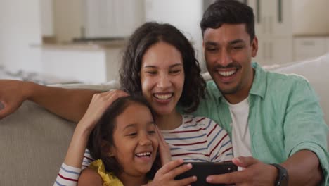 Happy-hispanic-parents-and-daughter-sitting-on-sofa-taking-selfie