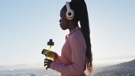Mujer-Afroamericana-Haciendo-Ejercicio-Al-Aire-Libre-Usando-Auriculares-Inalámbricos-Agua-Potable-En-El-Campo