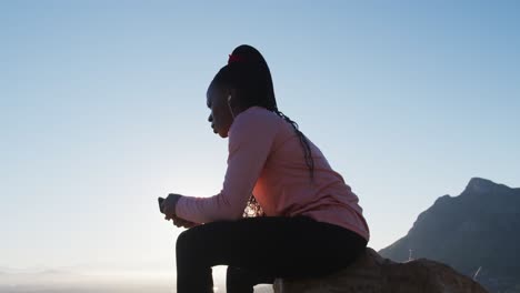 African-american-woman-exercising-outdoors-sitting-on-rock-using-smartphone-in-countryside-at-sunset