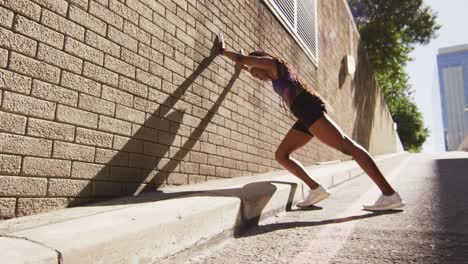 African-american-woman-exercising-outdoors-leaning-of-wall-and-stretching-in-the-city