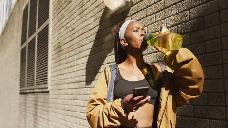 Mujer-Afroamericana-Haciendo-Ejercicio-Al-Aire-Libre-Bebiendo-Agua-Y-Usando-Un-Teléfono-Inteligente-En-La-Ciudad
