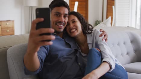Happy-hispanic-couple-embracing-on-sofa-in-living-room-taking-selfie