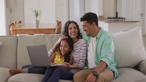 Happy-hispanic-parents-and-daughter-sitting-on-sofa-using-laptop