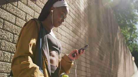 Mujer-Afroamericana-Haciendo-Ejercicio-Al-Aire-Libre-Usando-Auriculares-Usando-Un-Teléfono-Inteligente-En-La-Ciudad