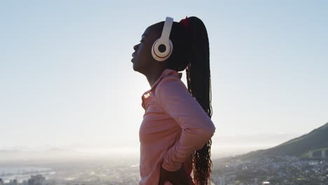 African-american-woman-exercising-outdoors-putting-on-wireless-headphone-in-countryside-at-sunset