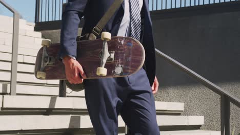 Midsection-of-caucasian-businessman-walking-downstairs,-holding-skateboard-on-sunny-day