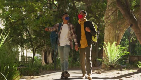 Two-happy-mixed-race-gay-male-couple-walking-and-talking-in-park-with-backpacks
