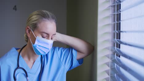 Portrait-of-caucasian-female-doctor-wearing-face-mask-looking-through-the-window-in-hospital-room