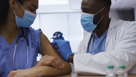 African-american-male-doctor-giving-covid-vaccination-to-female-colleague,-both-in-face-masks