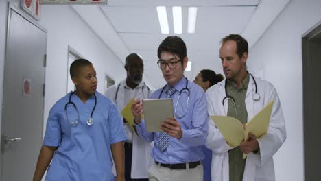 Diverse-group-of-male-and-female-doctors-talking-in-hospital-corridor,-walking-with-tablet-and-file