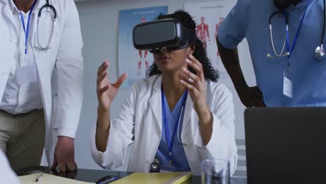 Asian-female-doctor-at-table-using-vr-headset-with-a-diverse-group-of-colleagues-gathered-around-her