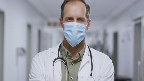 Portrait-of-caucasian-male-doctor-with-arms-crossed-wearing-face-mask-in-the-corridor-at-hospital