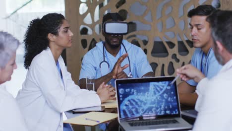 Diverse-group-of-doctors-sitting-in-discussion-at-a-meeting-using-laptop-and-vr-headset