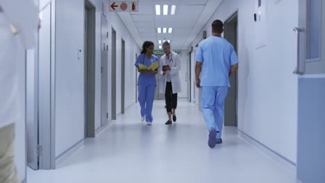 Two-diverse-female-doctors-walking-in-busy-hospital-corridor,-looking-at-tablet-and-file-and-talking
