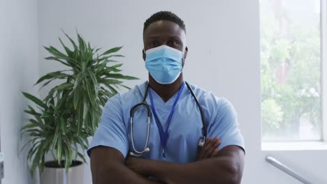 Portrait-of-african-american-male-doctor-wearing-scrubs-and-face-mask,-crossing-arms-in-hospital