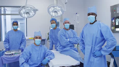 Diverse-male-and-female-doctors-wearing-face-masks-standing-in-operating-theatre-smiling-to-camera