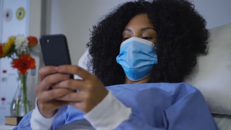 African-american-female-patient-wearing-face-mask-lying-on-hospital-bed-using-smartphone