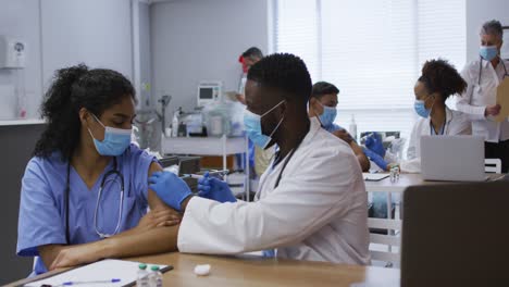 African-american-male-doctor-giving-covid-vaccination-to-female-colleague,-both-in-face-masks