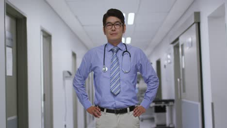 Porträt-Eines-Asiatischen-Männlichen-Arztes-Mit-Den-Händen-Auf-Den-Hüften,-Der-Lächelt,-Während-Er-Auf-Dem-Flur-Im-Krankenhaus-Steht
