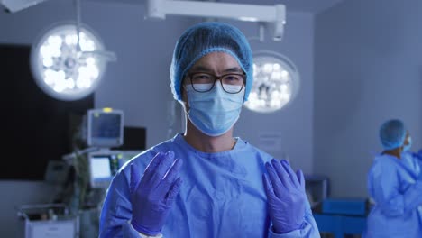 Portrait-of-asian-male-surgeon-wearing-face-mask,-gloves,-cap-and-scrubs-in-operating-theatre