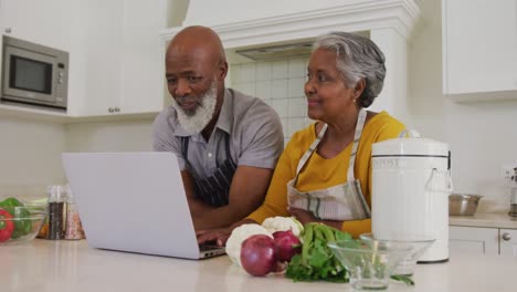 Afroamerikanisches-älteres-Paar-Trägt-Schürzen-Und-Benutzt-Gemeinsam-Einen-Laptop-In-Der-Küche-Zu-Hause